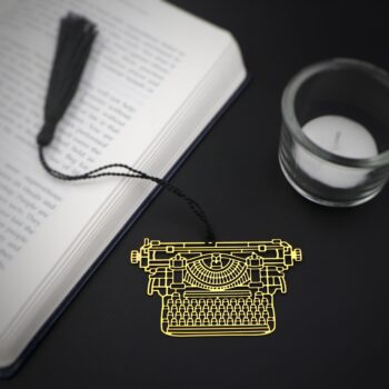 A close up of a metal bookmark shaped like Vintage Typewriter, delicately placed atop black background with black tassel on the book.